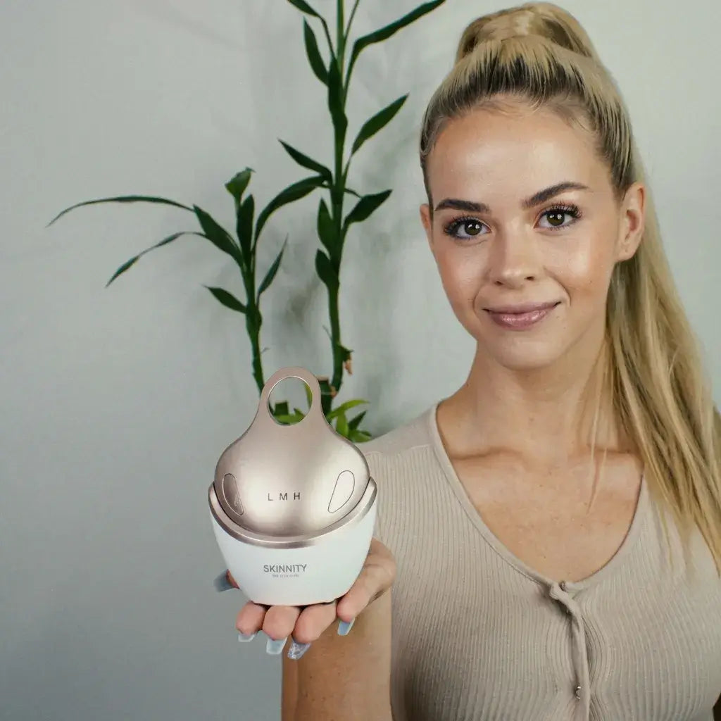 A woman holds the Quadrax Beauty Device in front of green plants, showcasing its sleek design and functionality.