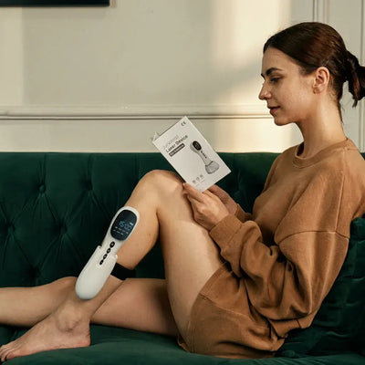 Woman relaxing on a couch using Dernox Pro Cold Laser + TENS on her leg while reading an instructions manual.