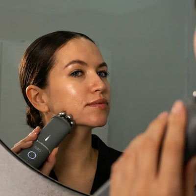 Woman using Vellura-Gess Contour & Renew device in front of a mirror for skincare.